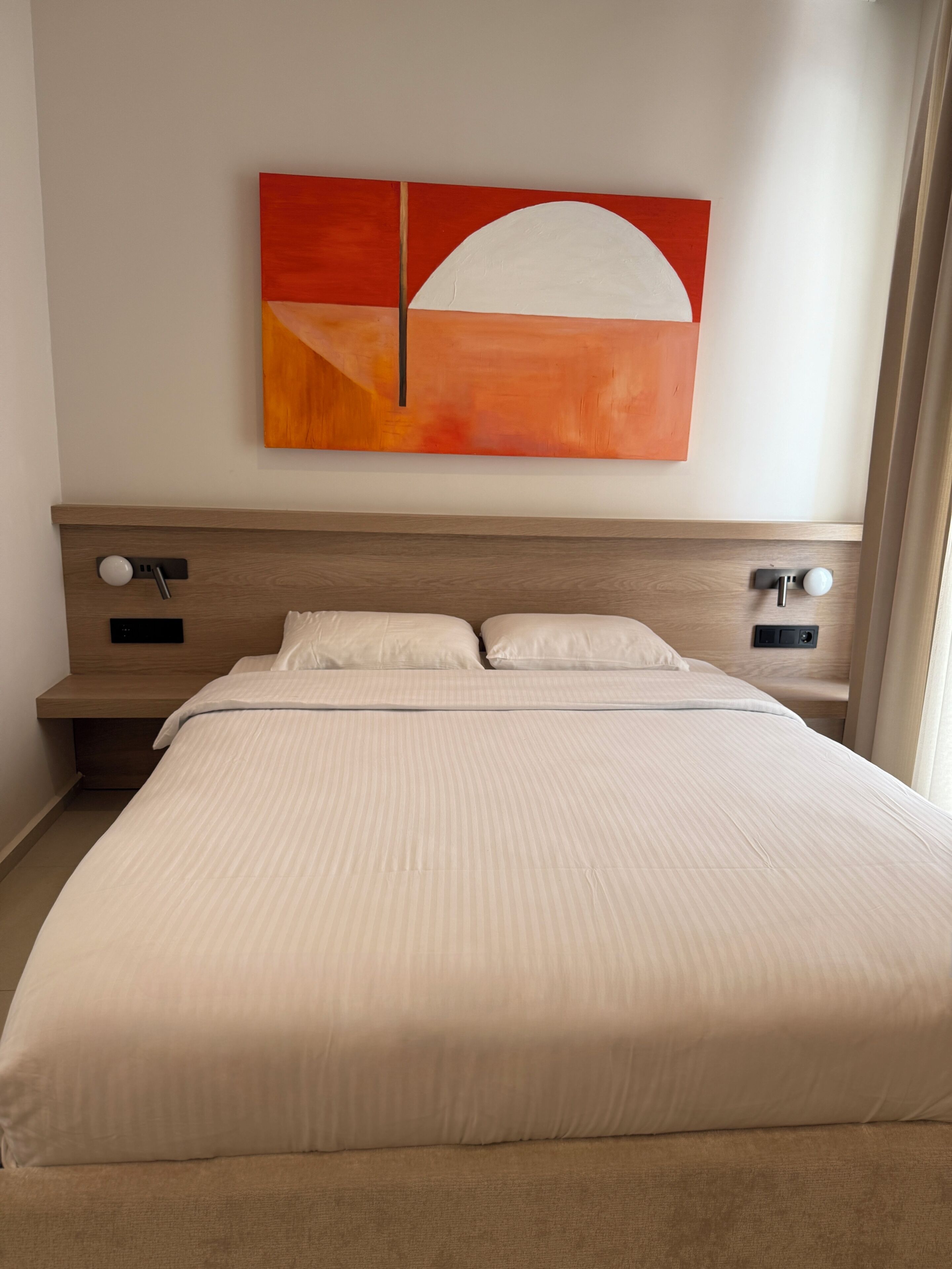 A hotel room with a large bed, two bedside tables, a wall-mounted artwork, and a window with curtains.