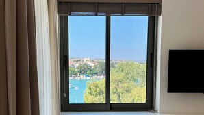 A room with a large window overlooking a coastal town, a wooden chair, and a flat-screen TV mounted on the wall. - Datca Deniz Hotel (Datça)