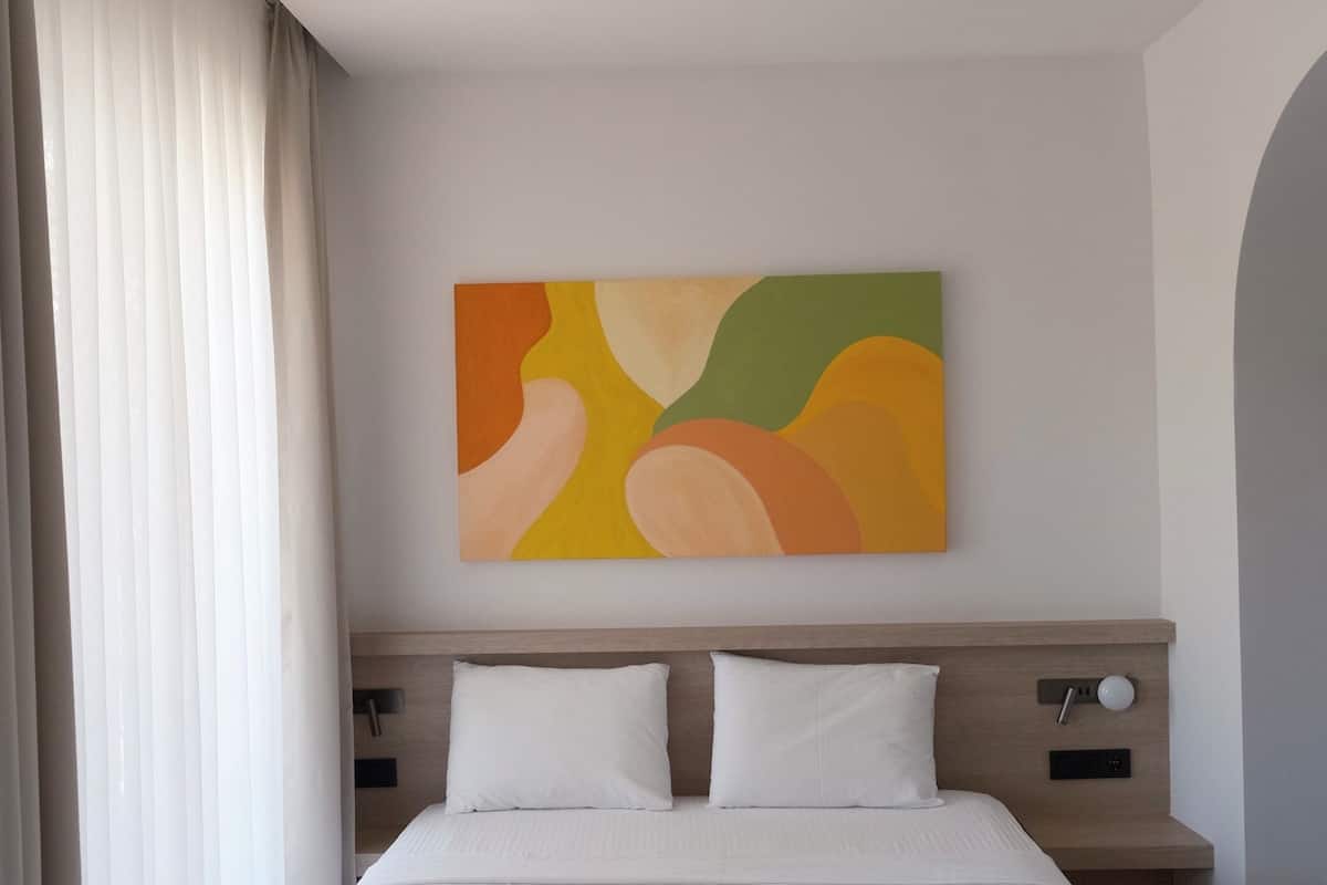 A modern hotel room with a large bed, a headboard with a control panel, a bedside table, and a colorful abstract painting on the wall.