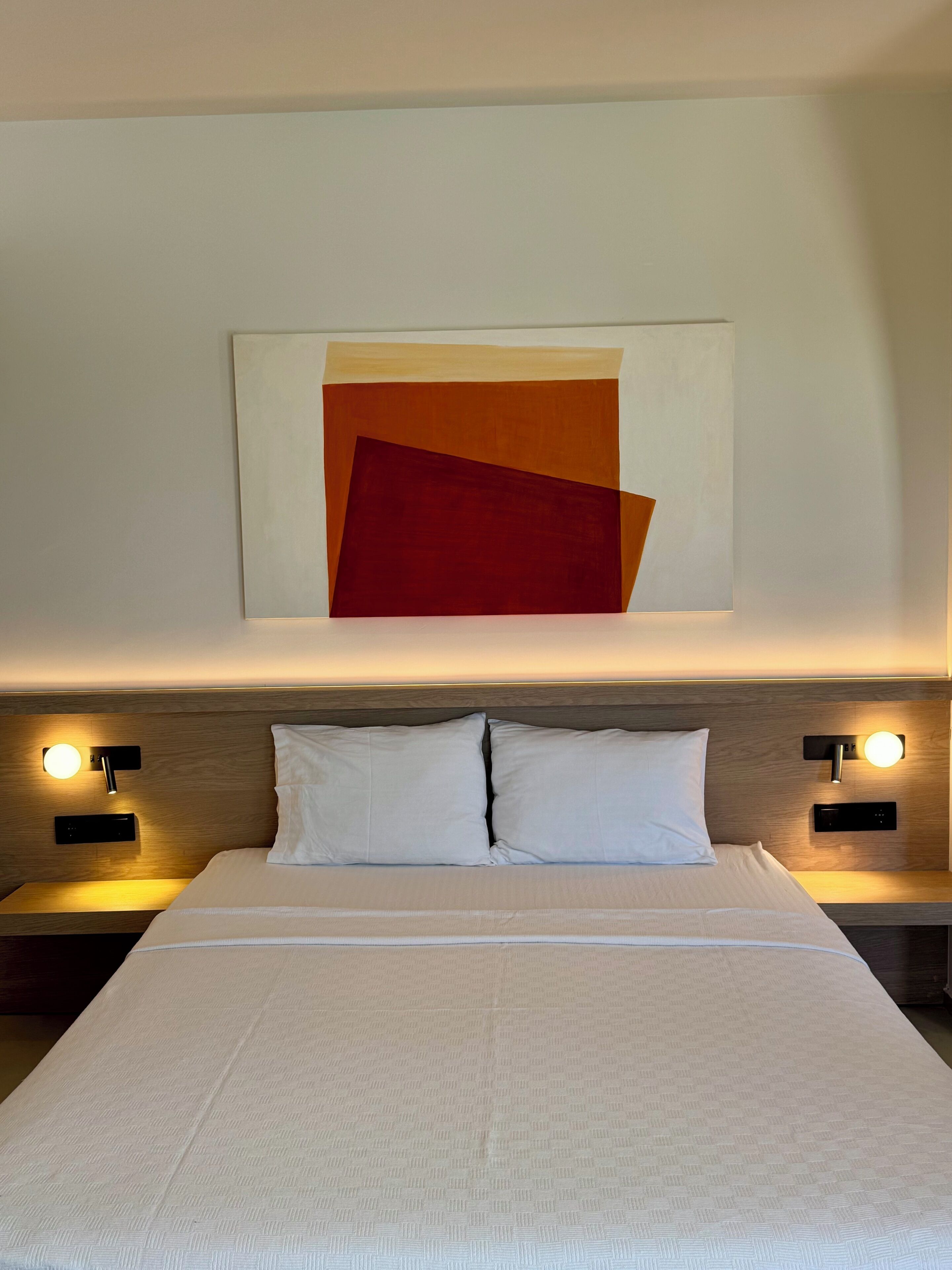 Ein Hotelzimmer mit einem großen Bett, zwei Nachttischen mit Lampen und einem an der Wand hängenden, abstrakten Gemälde.