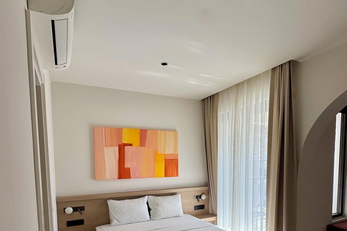 A modern hotel room with a large bed, a bedside table, a nightstand, a wall-mounted air conditioning unit, and a window with curtains.