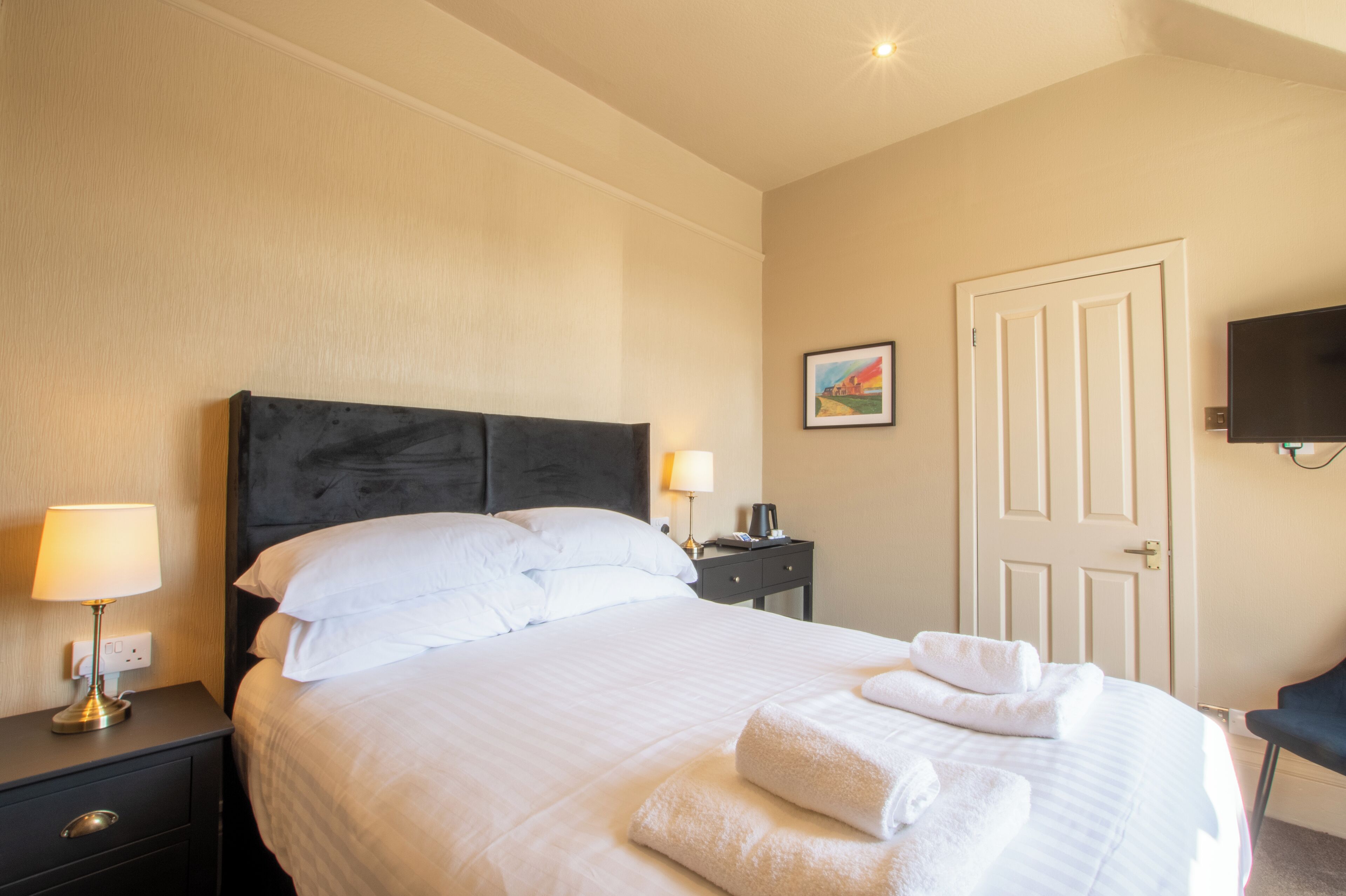 Quarto de hotel com cama, mesa de cabeceira com luminária, escrivaninha também com luminária, uma cadeira e uma porta.