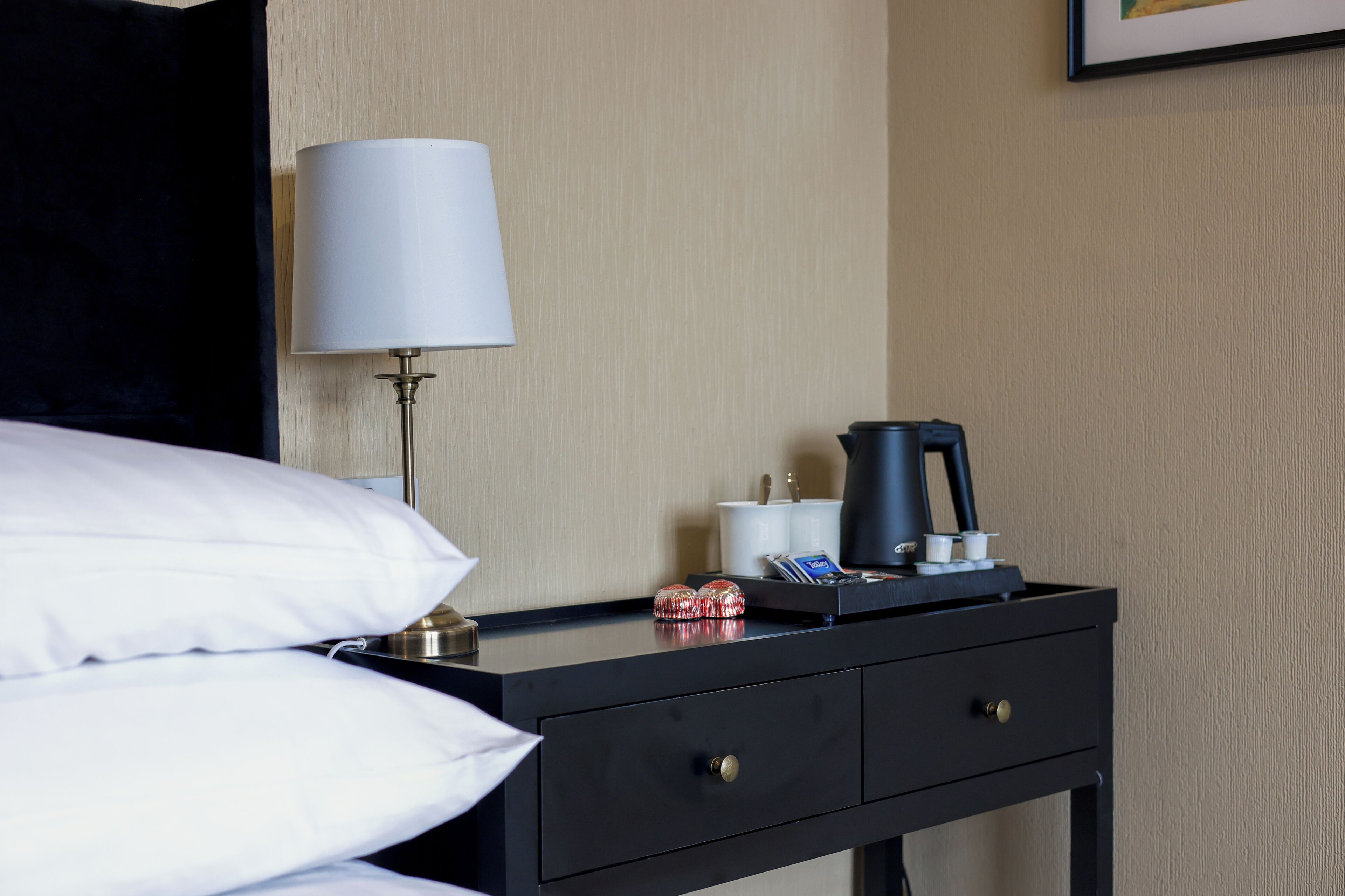 Habitación de hotel impecable con cama, lámpara de noche, cafetera y una mesita con tazas y una tarjeta.