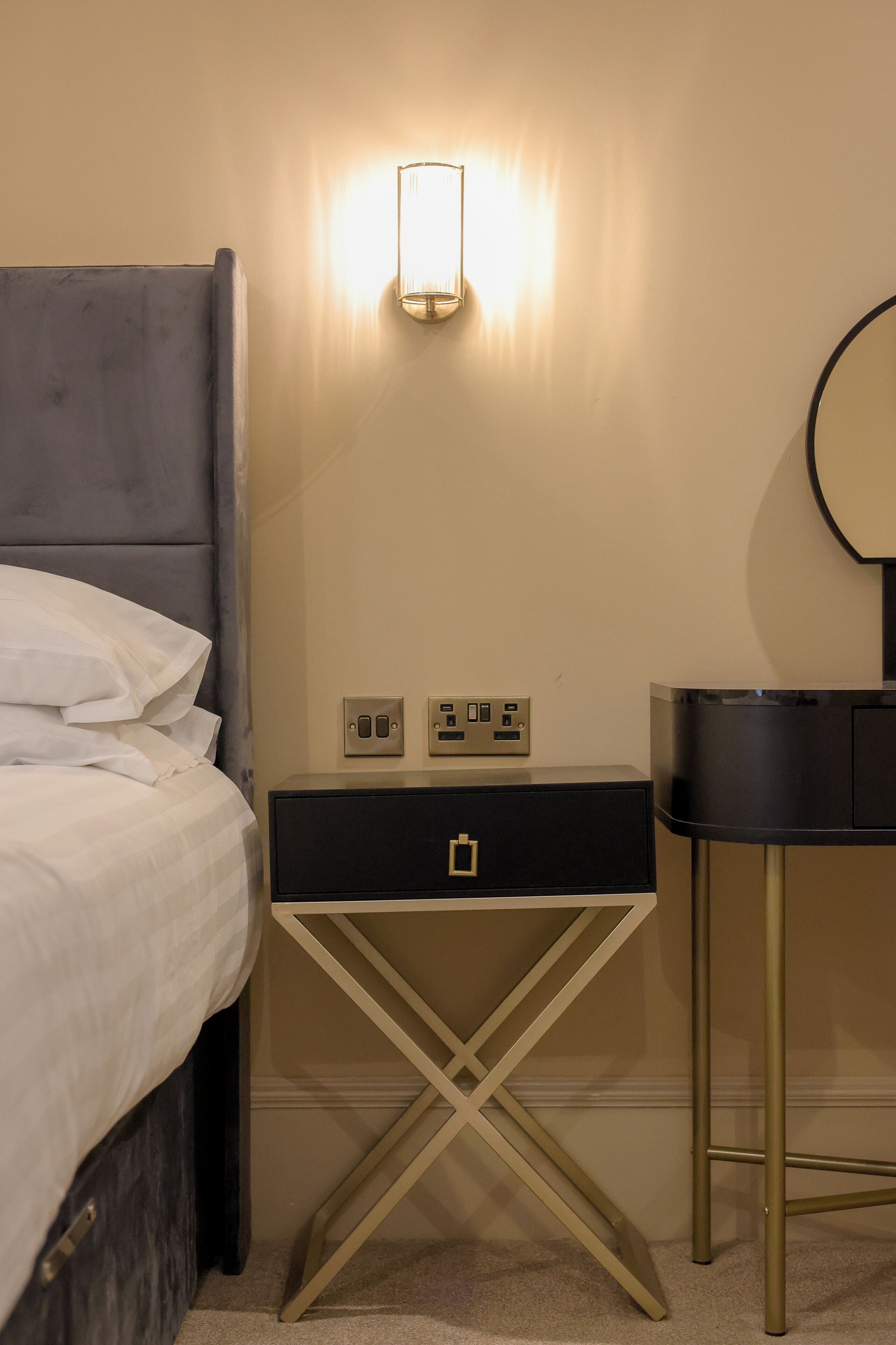 Un lit bien fait, avec du linge de lit blanc, une table de chevet avec une lampe et un petit miroir accroché au mur.