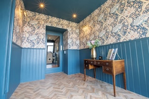 A room with blue walls, a herringbone wooden floor, and a patterned wallpaper. There is a wooden side table with a vase and a framed picture.