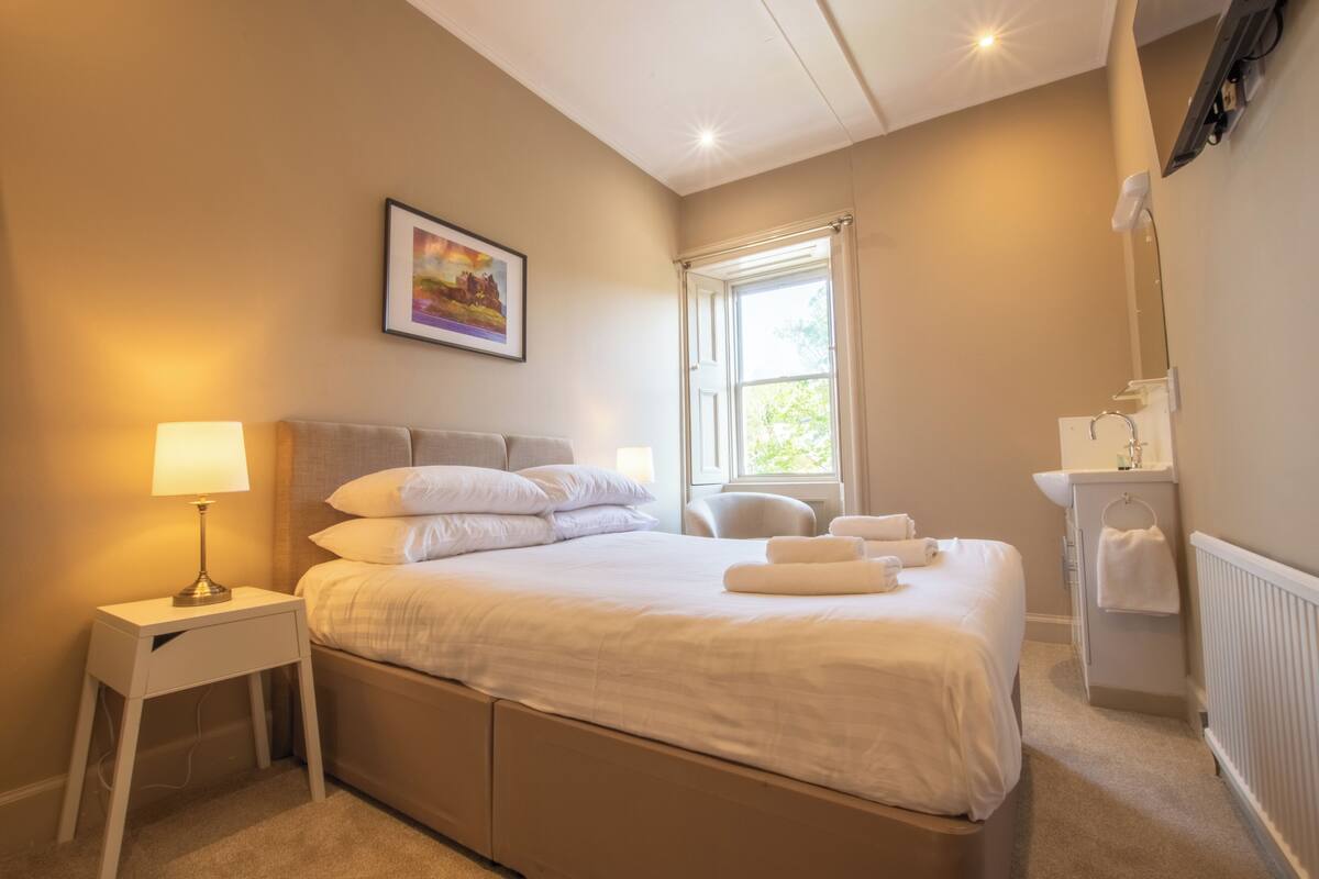 Un dormitorio ordenado con cama, mesita de noche, lámpara y ventana con cortinas.