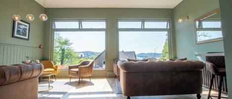 Ein geräumiges Wohnzimmer mit großen Fenstern, einem braunen Sofa, einem gelben Sessel und Holzdielen.