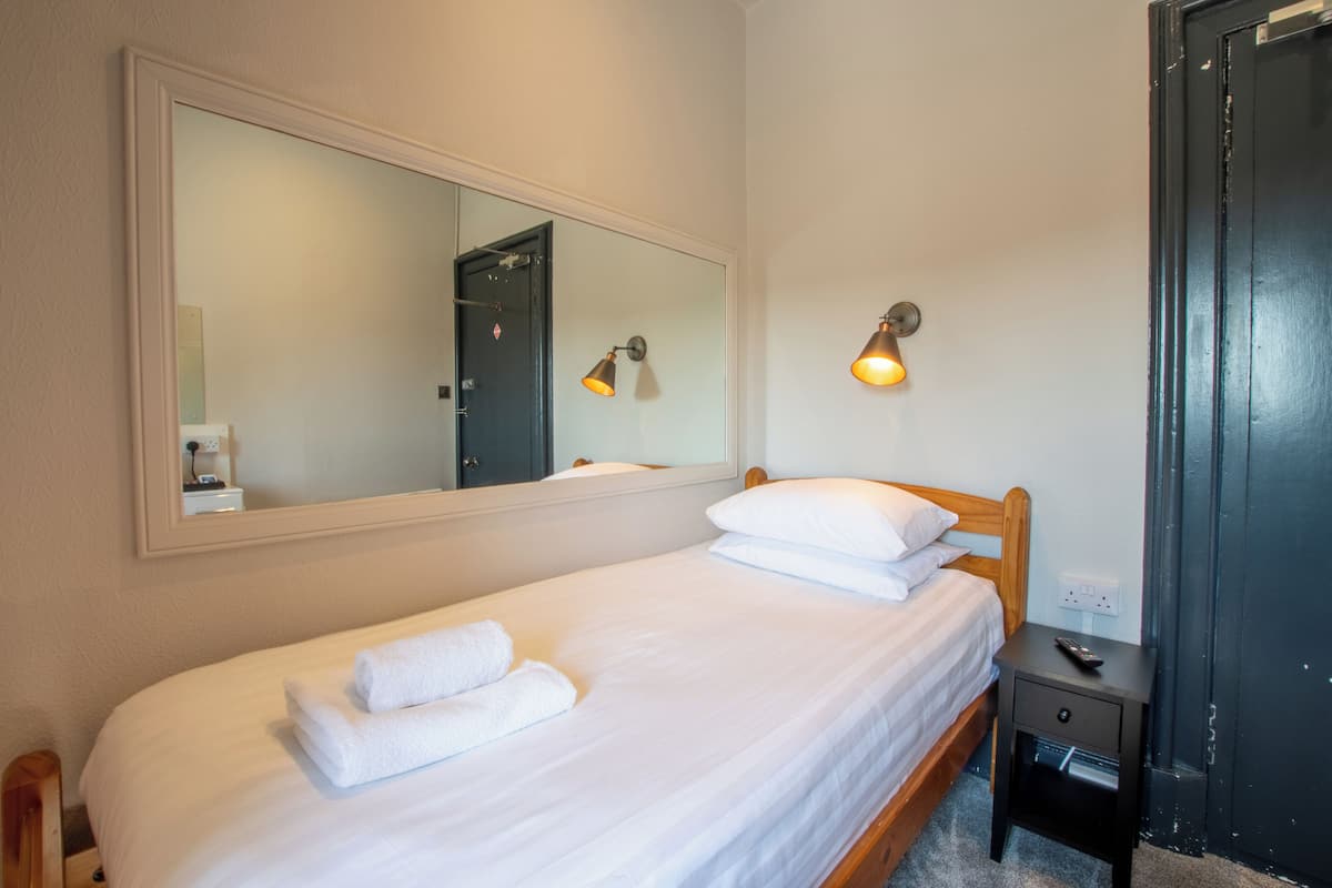 Una cama bien hecha con sábanas blancas y una toalla doblada, una mesita de noche con un control remoto y un espejo en la pared que refleja una puerta y una lámpara.