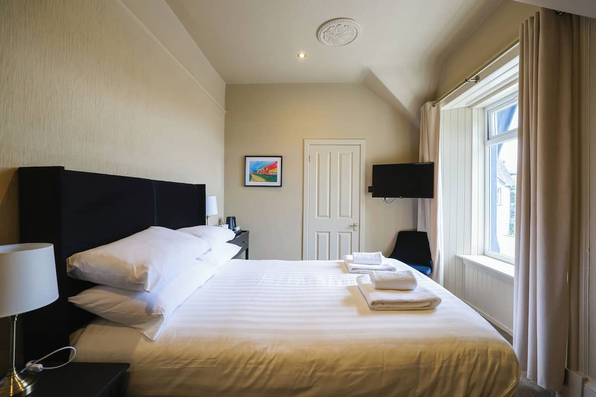 Una habitación de hotel con una cama grande, una mesita de noche con lámpara, un televisor de pantalla plana y una ventana con cortinas.