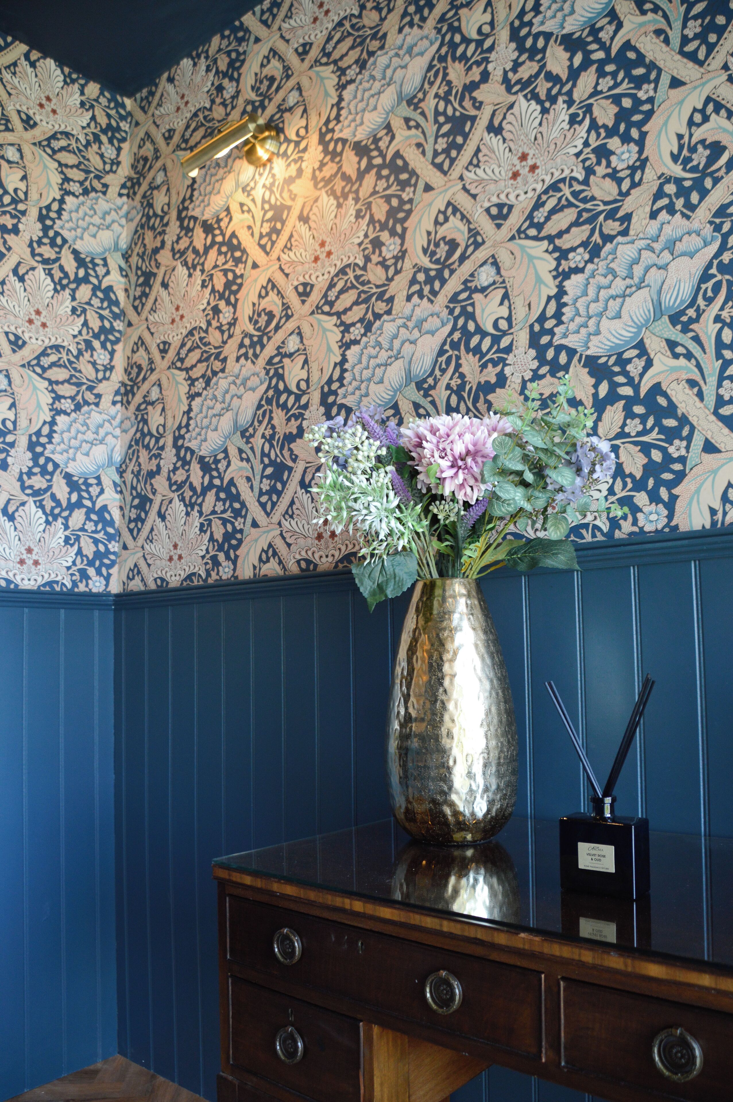 A decorative vase with flowers on a wooden dresser against a wall with a floral pattern.
