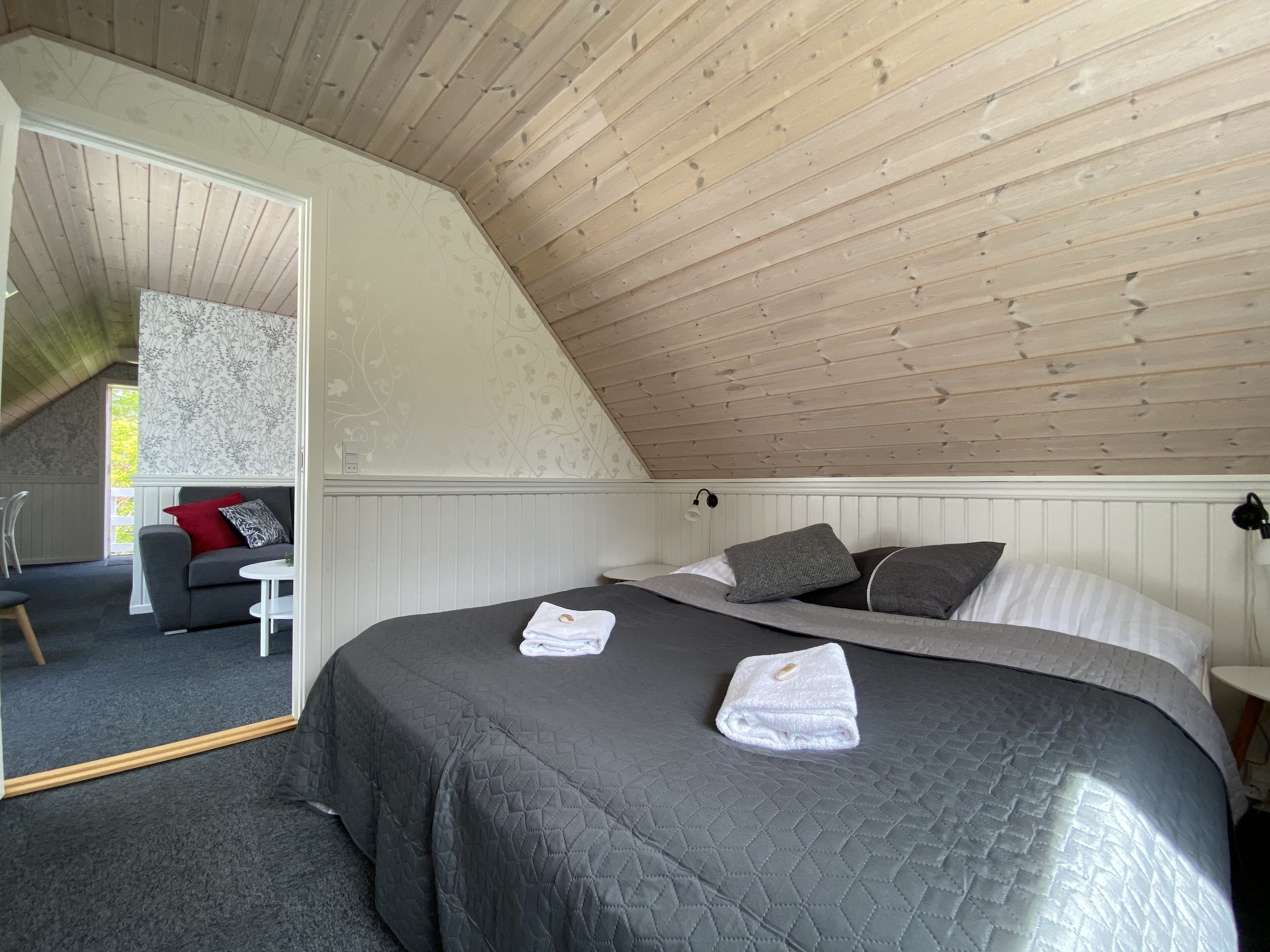 Une chambre avec un plafond en pente, un lit recouvert d’une couverture sombre, deux serviettes, une table de chevet et une chaise.