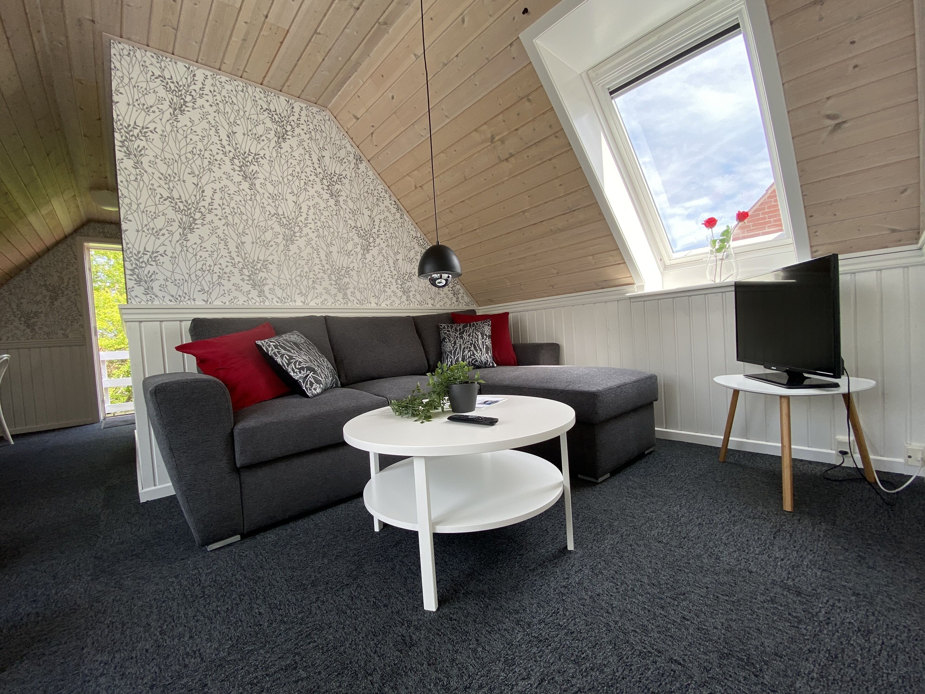 Un dormitorio tipo loft moderno con techo inclinado, un sofá gris con cojines rojos y estampados, una mesa de centro redonda blanca y un televisor sobre un mueble de madera.