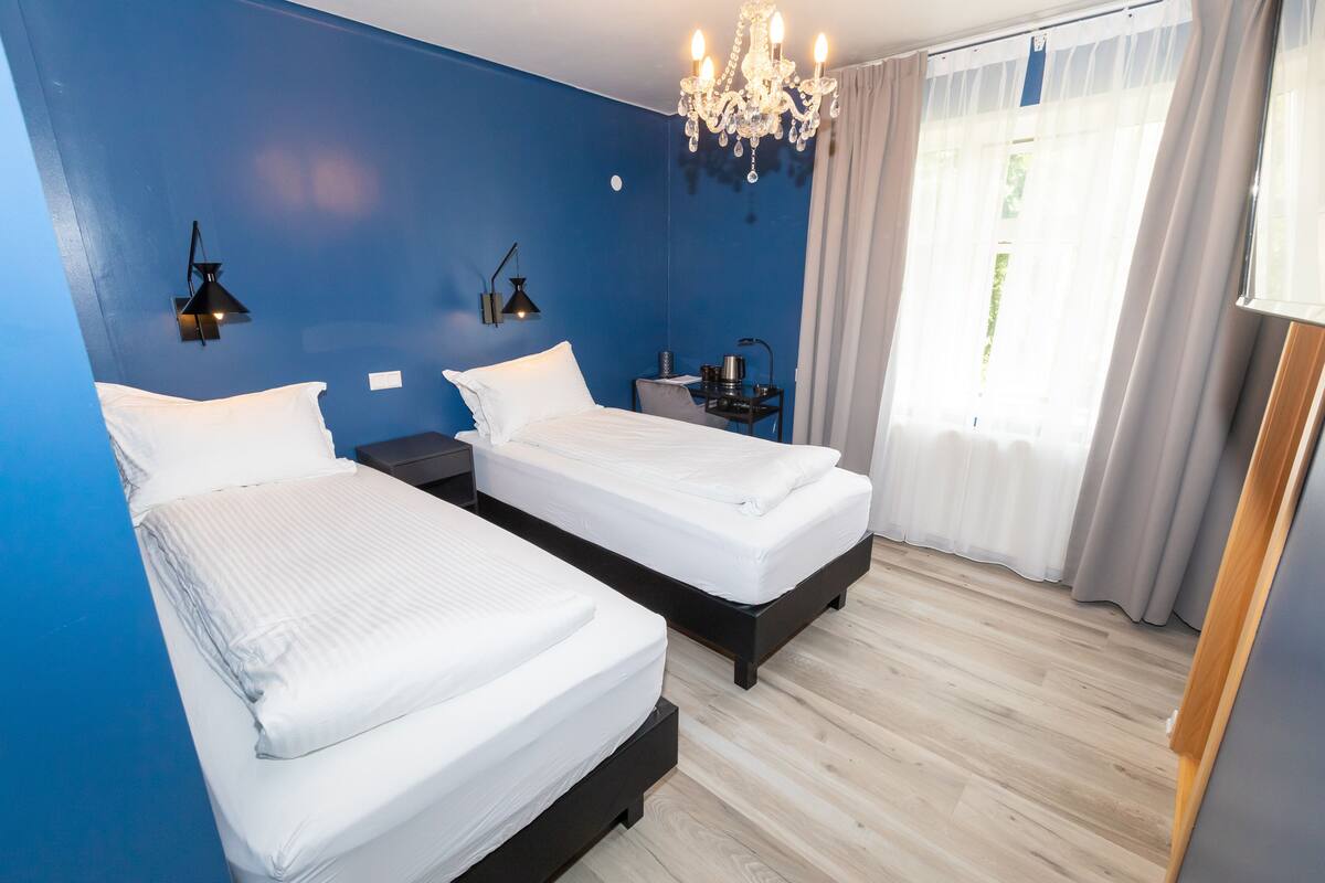 Una habitación de hotel con dos camas, una araña, una mesita de noche y una ventana con cortinas.