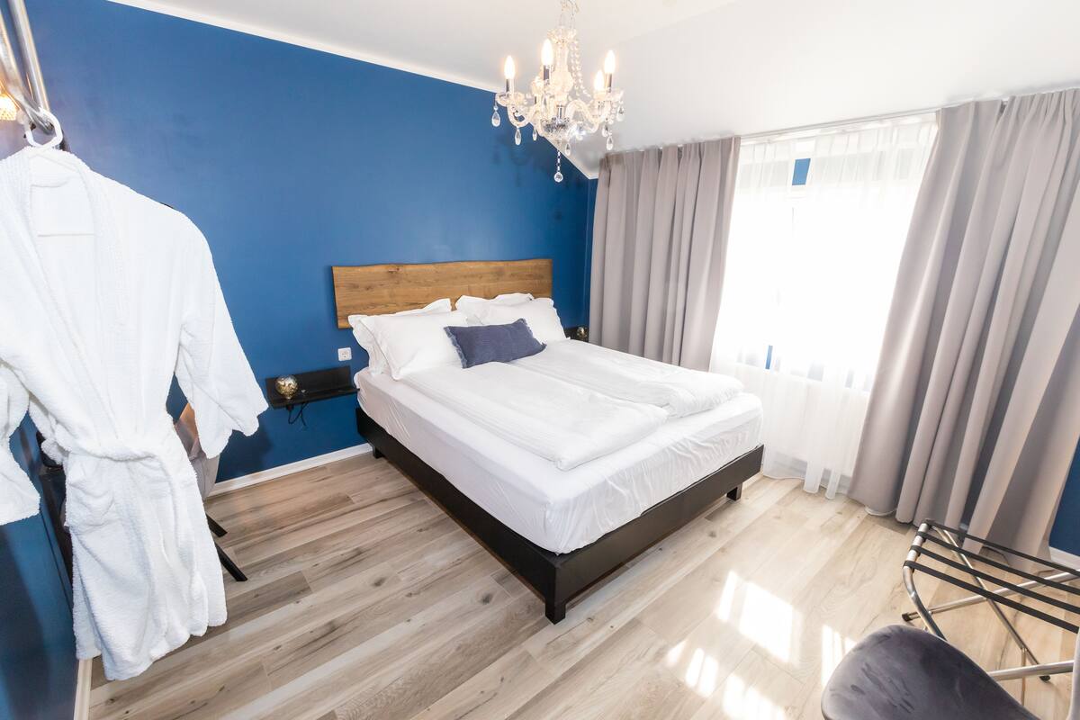 Un dormitorio con cabecera de madera, una cama blanca, una araña y vistas al exterior a través de una ventana con cortinas.