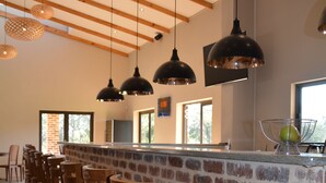 A bar area with wooden chairs, a brick counter, hanging pendant lights, and a view of the outdoors through large windows. - Esther's Country Lodge (Magaliesberg)