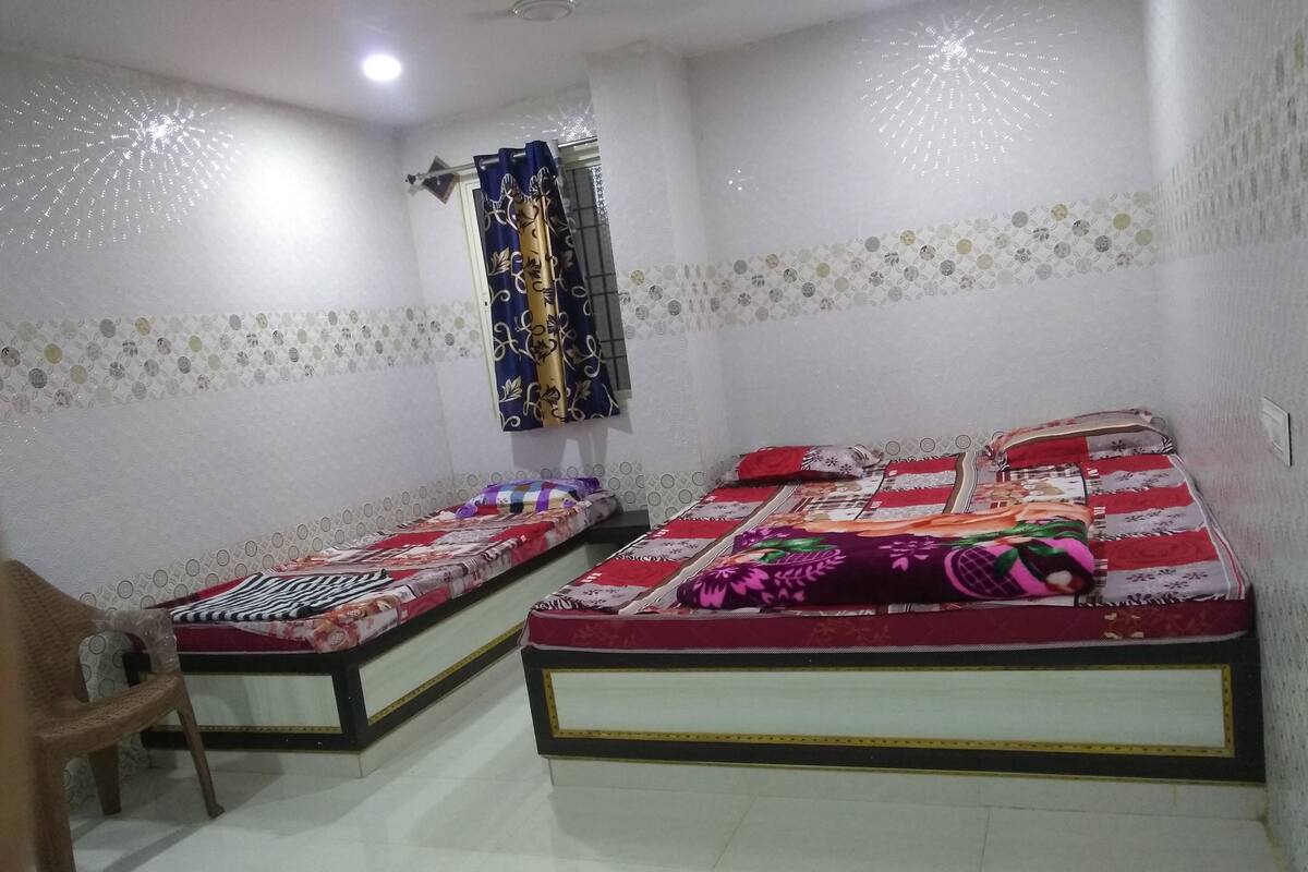 A small, well-lit room with two single beds, a chair, and a window with decorative curtains.