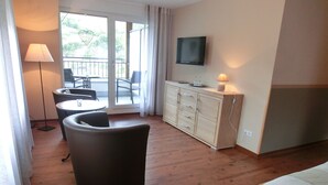 Une chambre d’hôtel comprenant un lit, un canapé, une table à manger avec des chaises, un téléviseur fixé au mur et un balcon donnant sur un espace verdoyant.