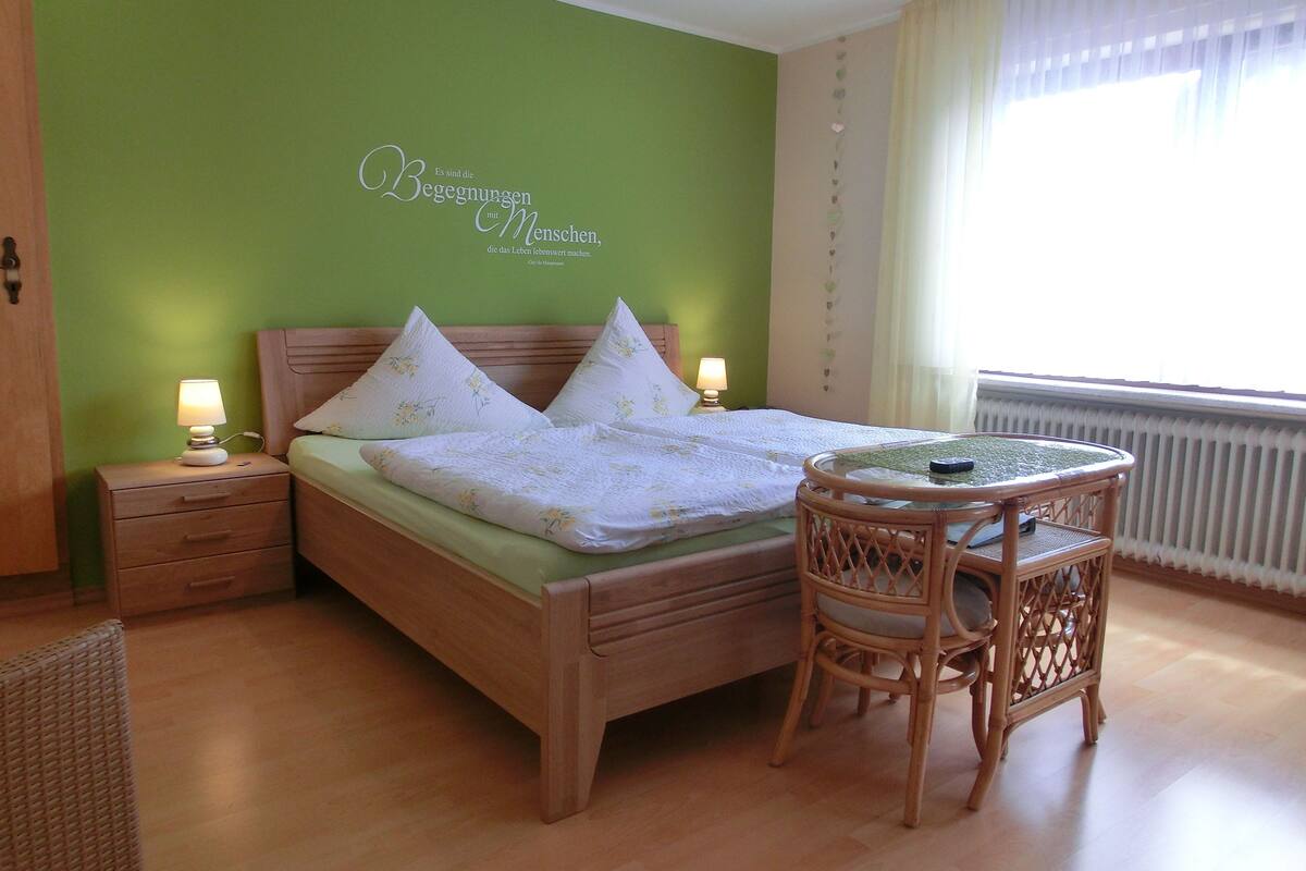 Un dormitorio ordenado con cama, mesitas de noche, una silla y una pequeña mesa.