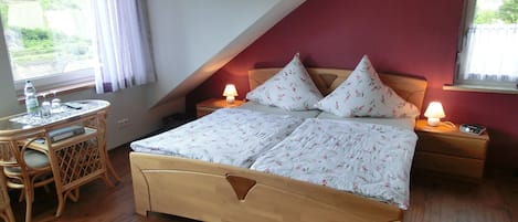 Quarto com cama de madeira, duas mesas de cabeceira com abajures, uma mesinha com uma cadeira e uma janela com cortinas.