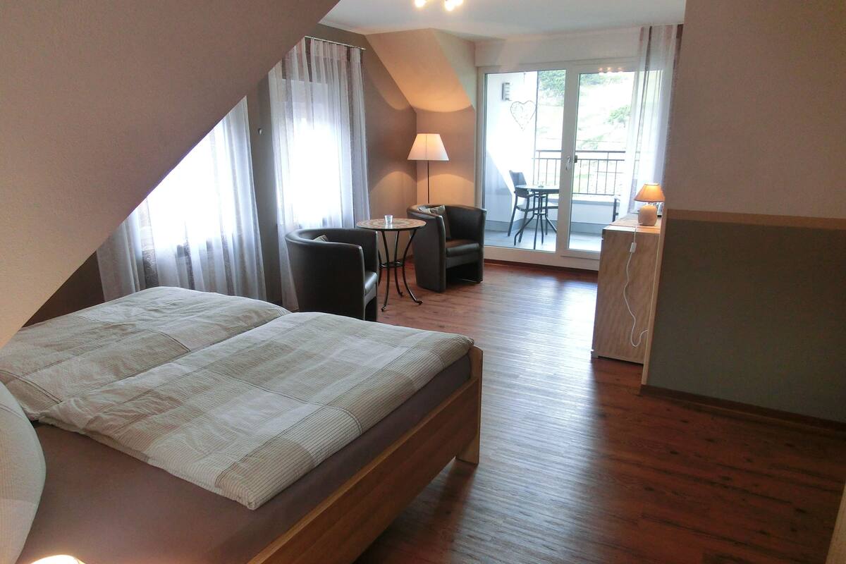 Un dormitorio con cama, escritorio y silla. Tiene un balcón con mesa y sillas.