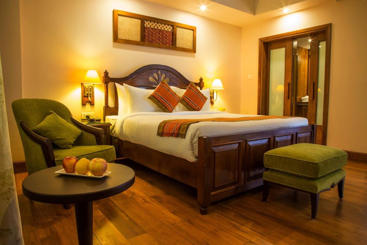 A hotel room with a wooden bed, green armchair, round table with apples, and a green ottoman.