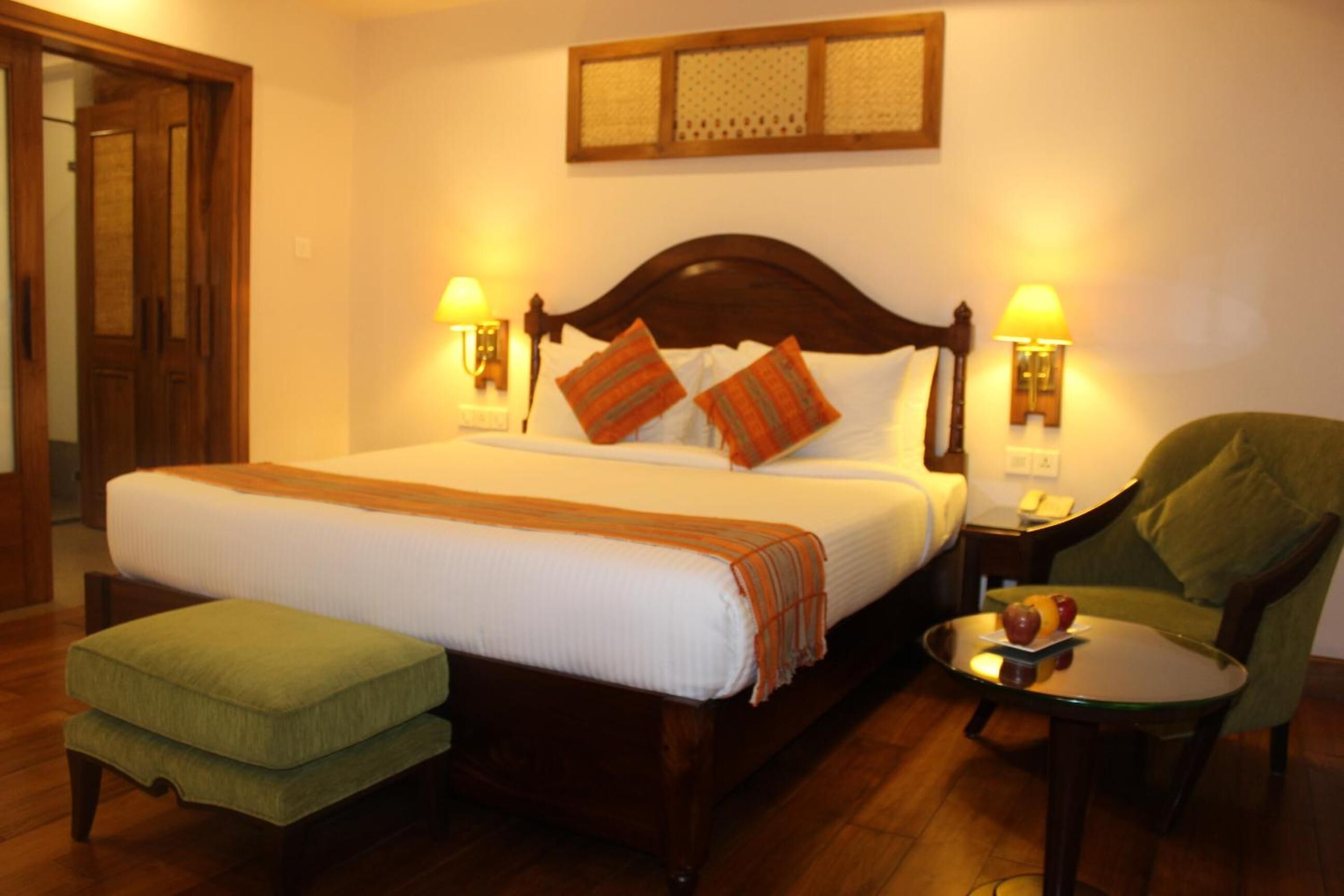 A neatly made bed with a wooden headboard, two bedside lamps, a green chair, and a small round table with fruits.