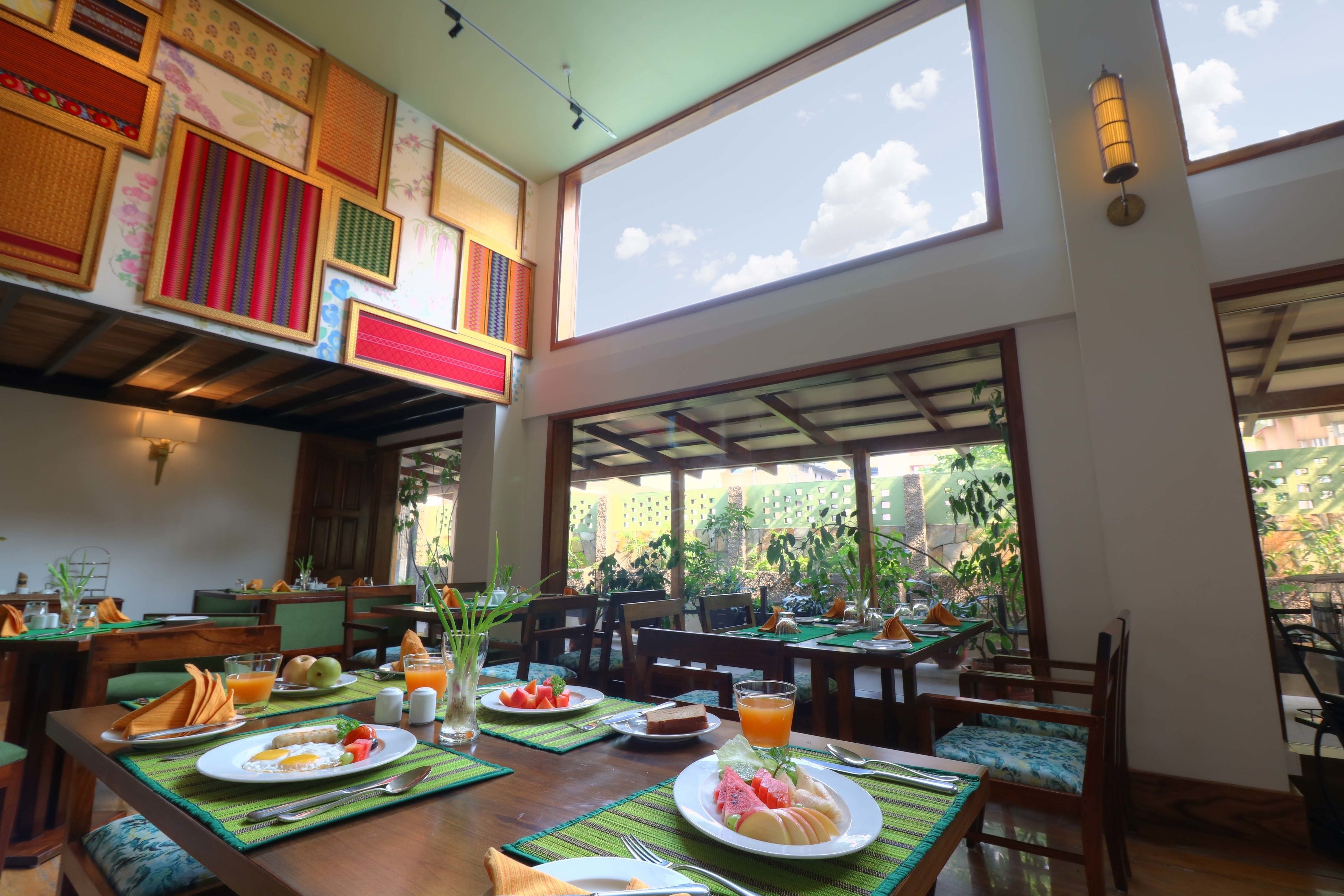 Un restaurante con mesas dispuestas para el desayuno, mostrando platos de comida, bebidas y un amplio ventanal con vistas a la vegetación.