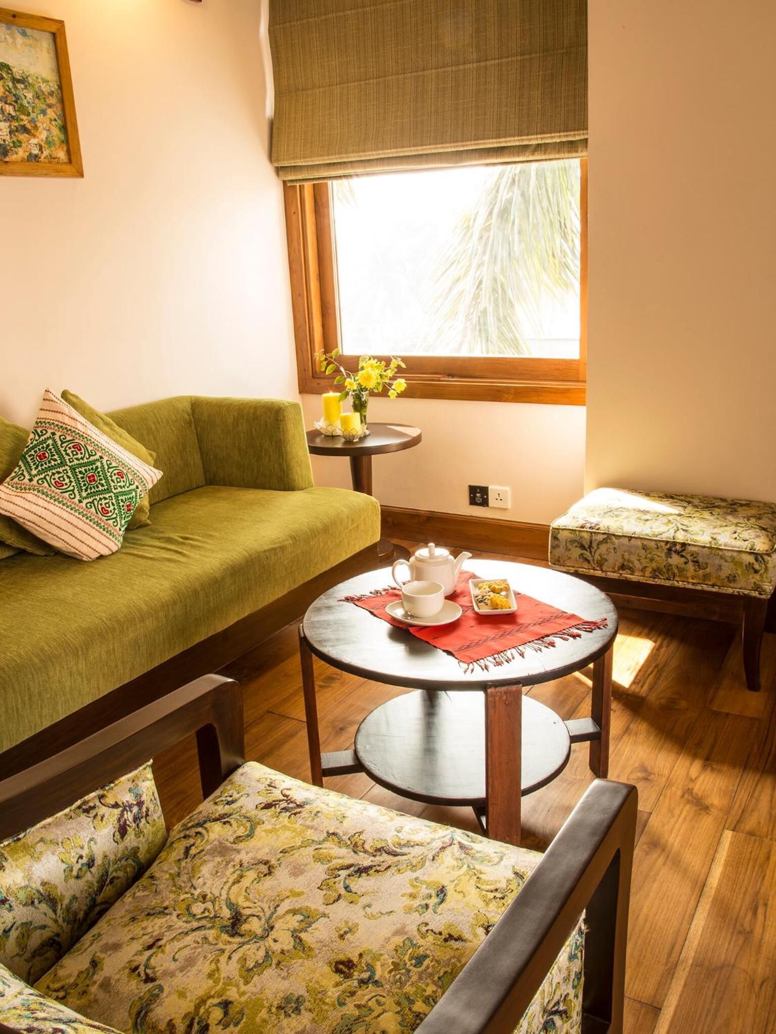 Un salon chaleureux avec un canapé vert, une table basse ronde sur laquelle se trouvent une théière et des tasses, et un pouf orné d’un motif floral.
