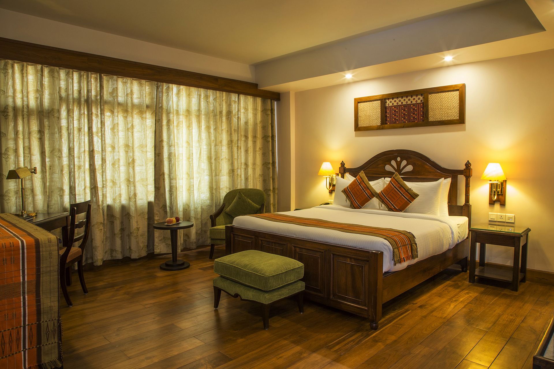 A hotel room with a wooden bed, two bedside lamps, a green chair, a small table with fruit, and a desk with a lamp.