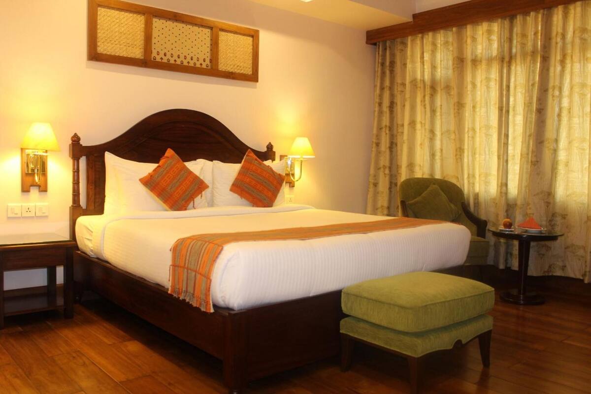 A hotel room with a wooden bed, two bedside lamps, a green armchair, a small table with a red object, and a window with curtains.