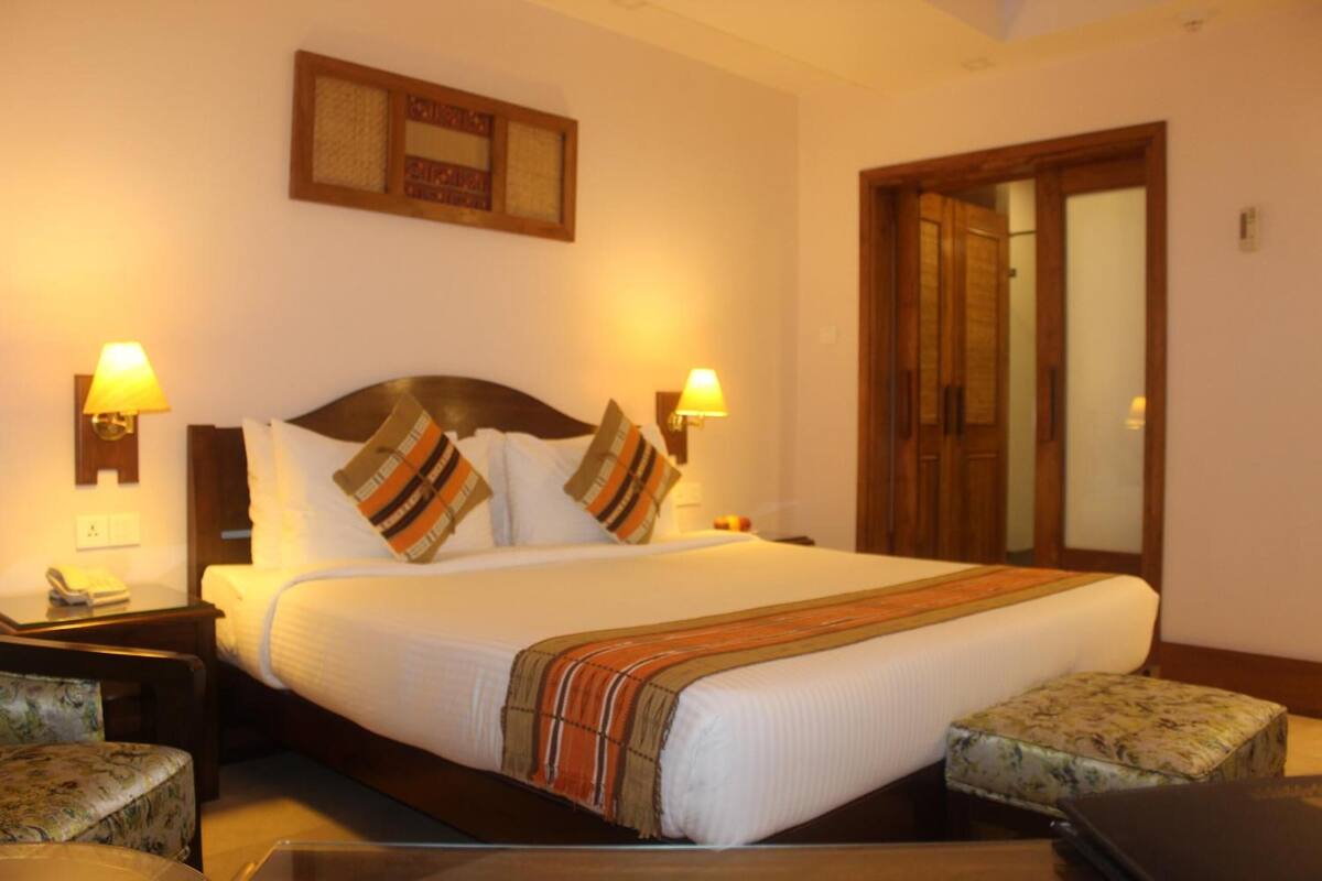 A neatly arranged hotel room with a large bed, two bedside lamps, a wooden headboard, a floral ottoman, and a mirror on the wall.