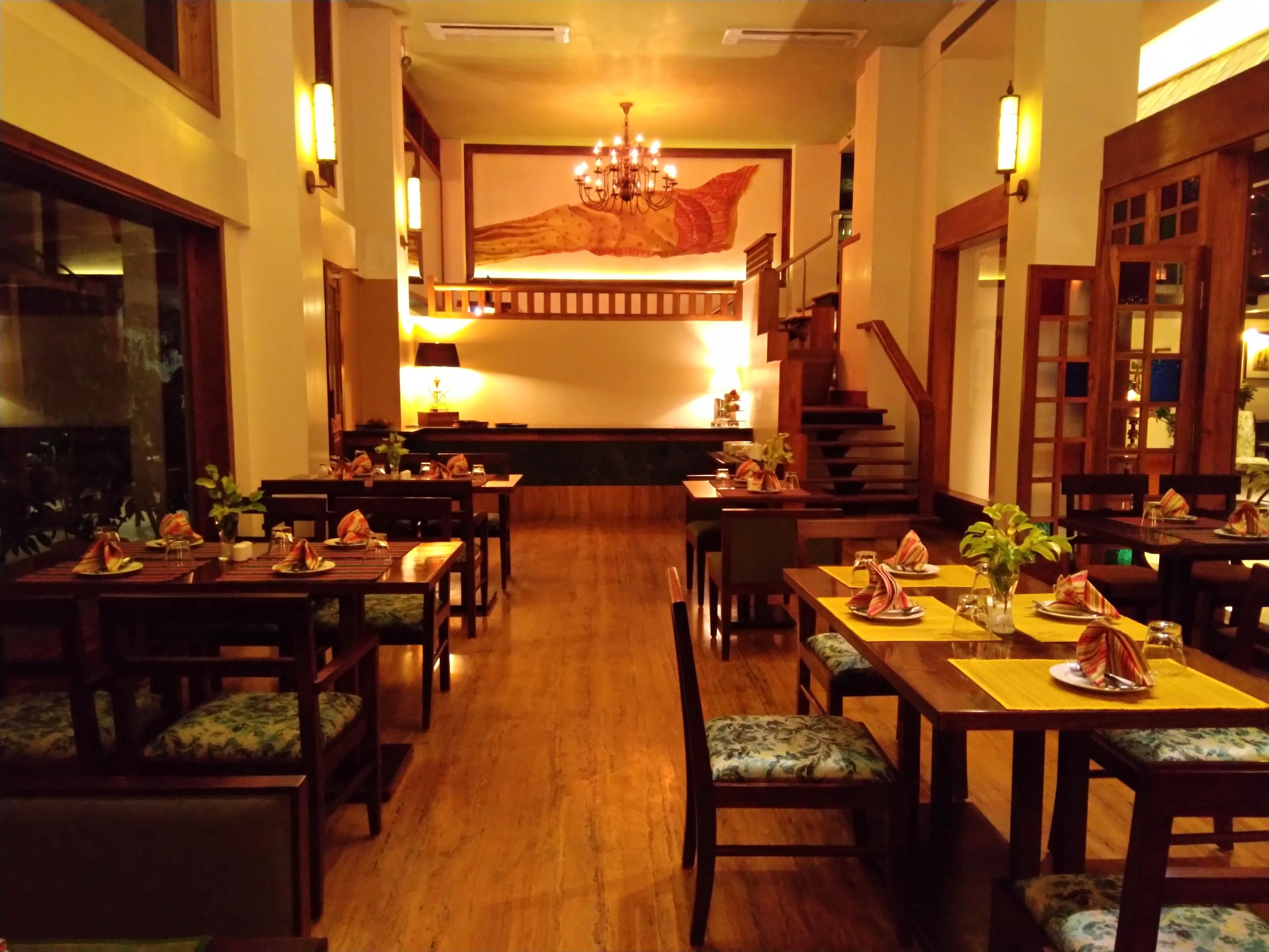 Un comedor amplio con mesas y bancos de madera, una escalera al fondo y un gran mural en la pared.