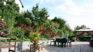Eine Terrasse im Freien mit einer Holzbank, einem Tisch und Stühlen. Übertöpfe mit Pflanzen und verschiedene Blumen sind ebenfalls vorhanden.