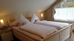 Ein Doppelbett mit Holzrahmen und weißer Bettwäsche, ein Nachttisch mit Lampe sowie ein Fenster mit Vorhängen.