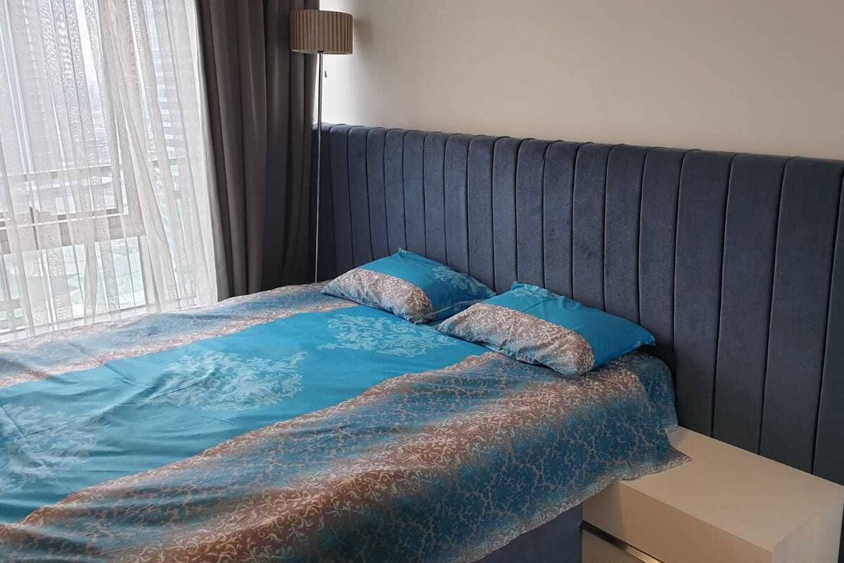 Une chambre à coucher avec une tête de lit sombre, un lit recouvert d’une literie à motifs bleus et une fenêtre ornée de rideaux légers.
