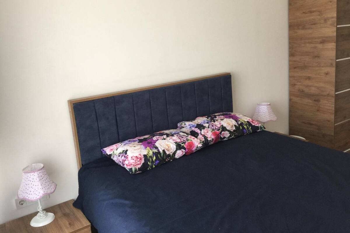 Un lit avec une tête de lit sombre et un coussin à motifs floraux, une lampe blanche avec un abat-jour rose et une armoire en bois.
