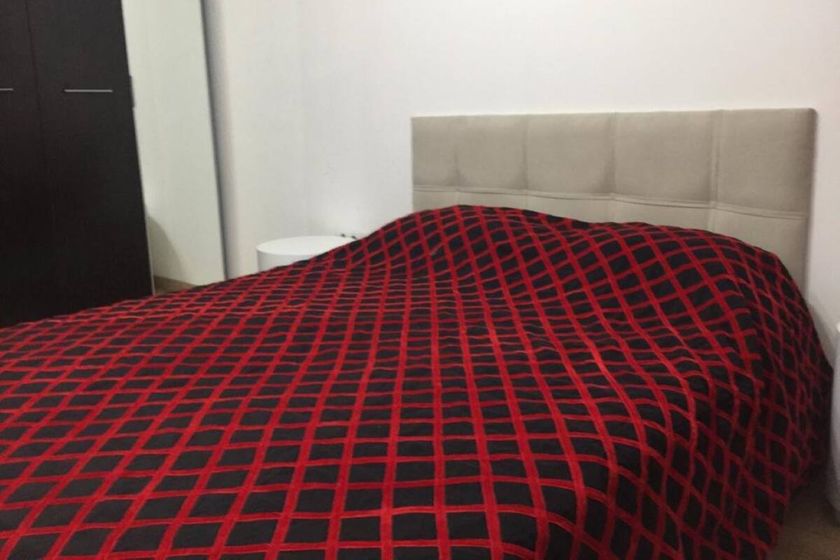 Un lit avec une couvre-lit à carreaux rouges et noirs, une tête de lit blanche et une table de chevet blanche sur le côté gauche.