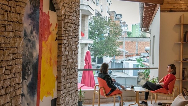 A spacious room with a stone wall, a large window, and wooden ceiling beams. - Mosaic Home - Hostel (Tirana)