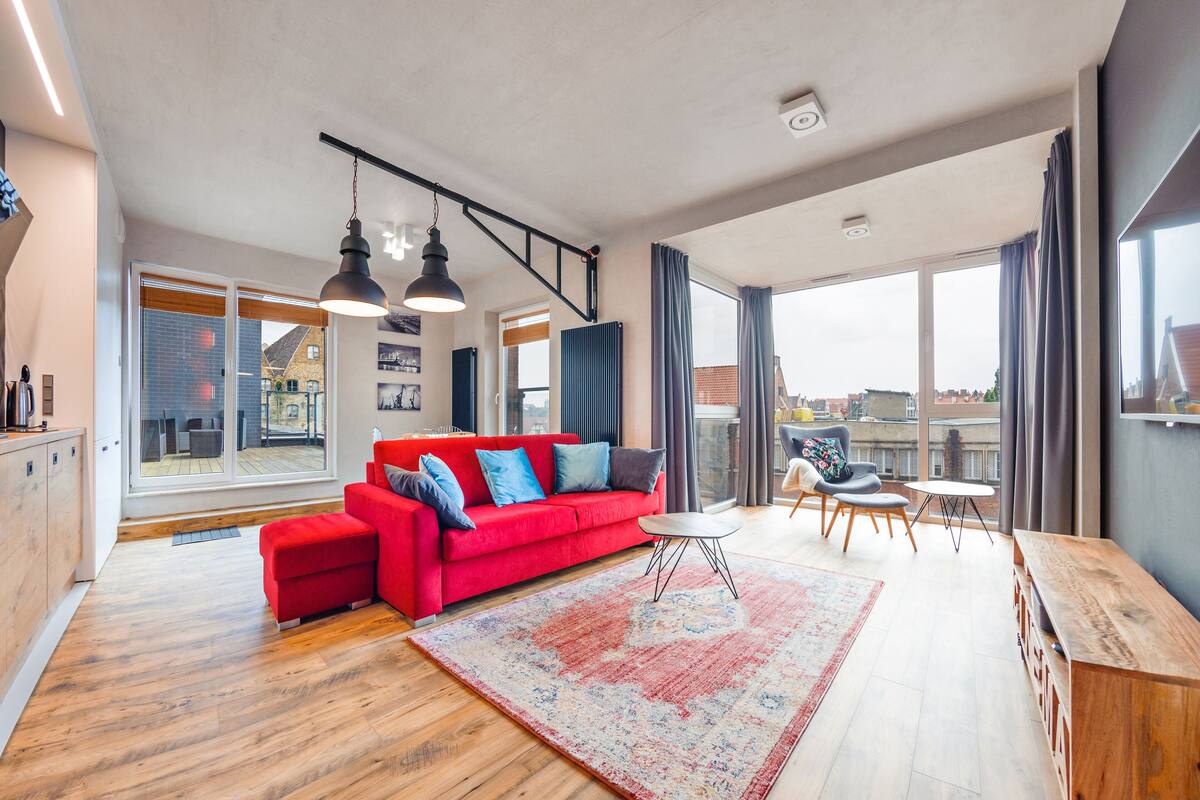 Un vaste espace de vie agrémenté d’un canapé rouge, de parquet et de grandes fenêtres offrant une vue sur la ville.