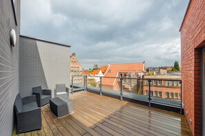 A rooftop terrace with wicker furniture, a glass railing, and a view of city buildings.