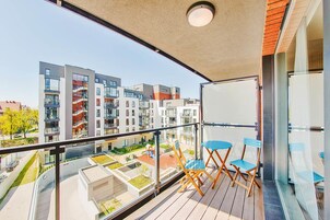 Un balcón con vistas a edificios de apartamentos modernos, barandilla de vidrio y sillas plegables azules.