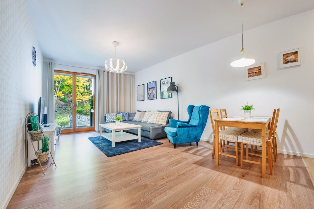 Ein modernes Wohnzimmer mit einer Couch, einem Esstisch mit Stühlen, einem Couchtisch und einer Deckenleuchte. An den Wänden befinden sich gerahmte Bilder und auf dem Tisch steht eine Pflanze.