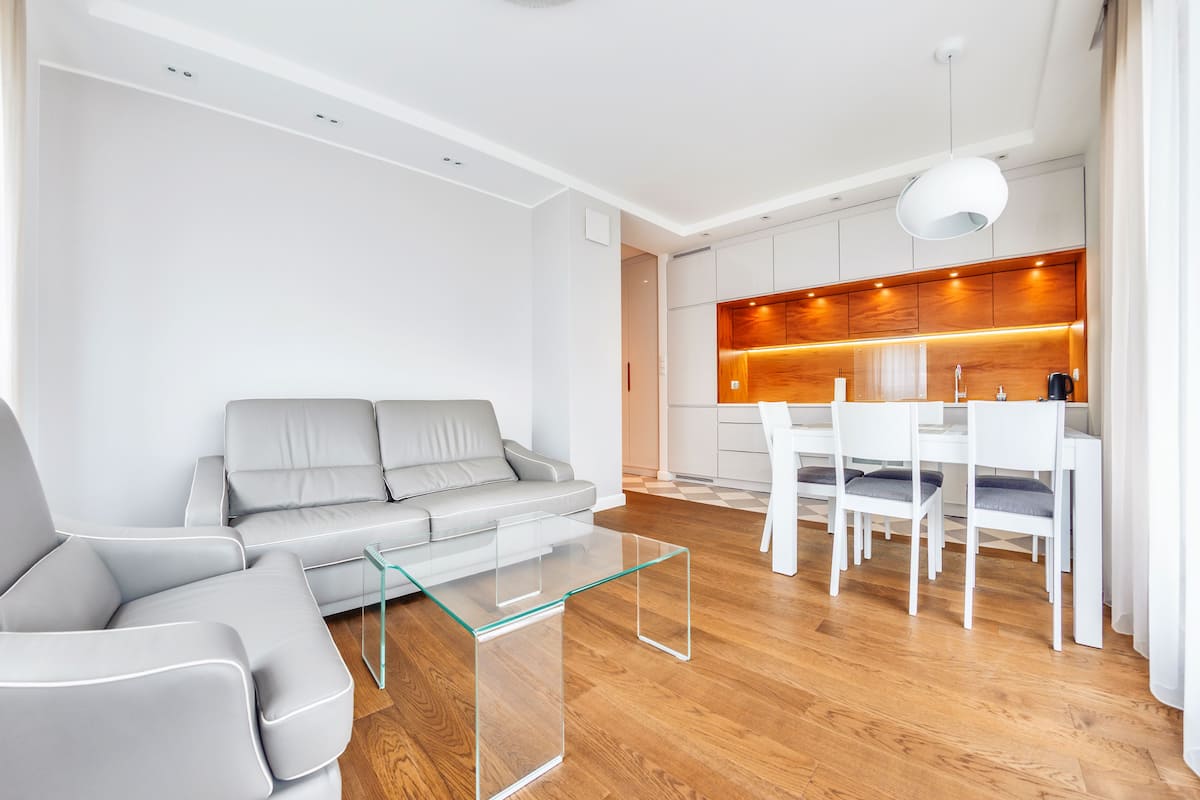Ein modernes Wohnzimmer mit einem grauen Sofa, einem Glastisch und einem Essbereich mit weißen Stühlen. Im Hintergrund ist eine Holzküche zu sehen.