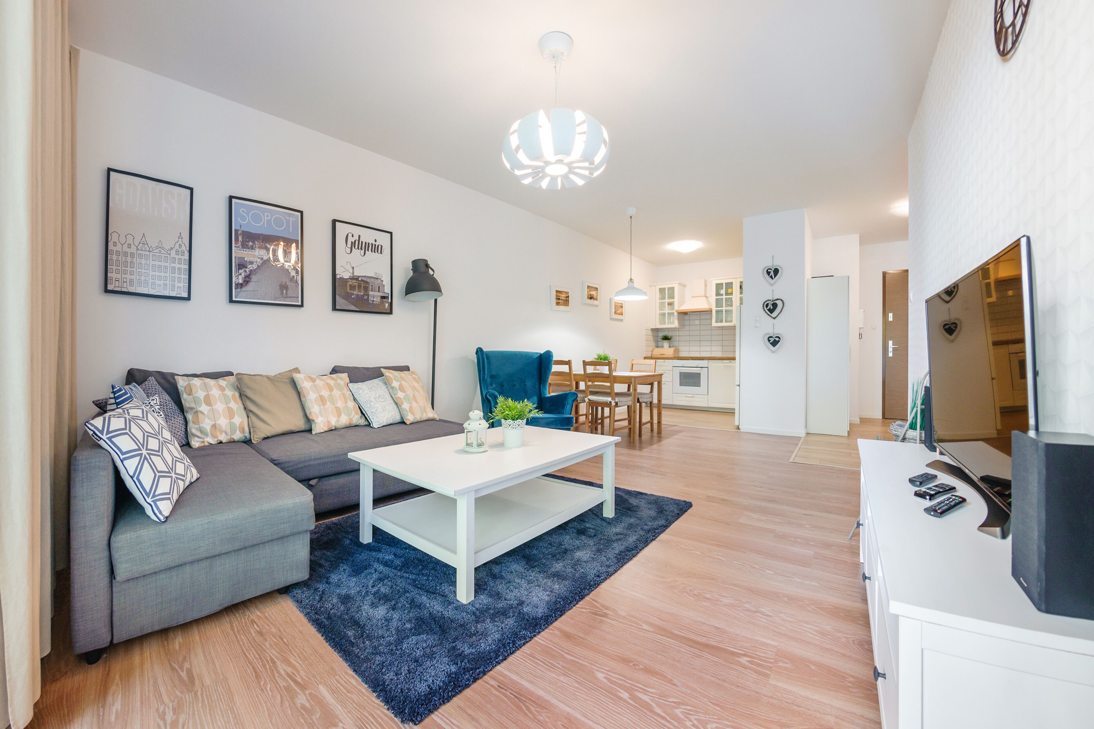 A modern living room with a grey sectional sofa, a white coffee table, and a blue rug. There are framed pictures on the wall, a kitchen in the background, and a dining area with a table and chairs.
