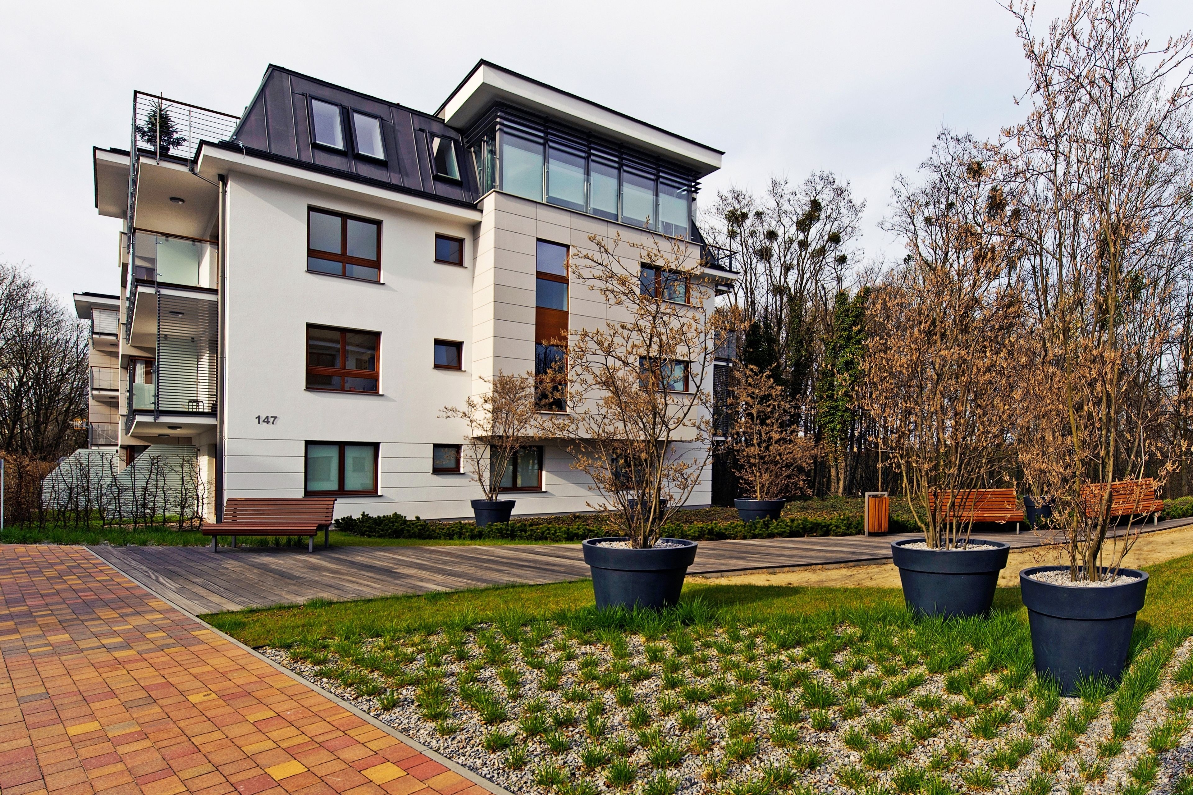 A modern multi-story residential building with a flat roof, large windows, and a paved driveway.