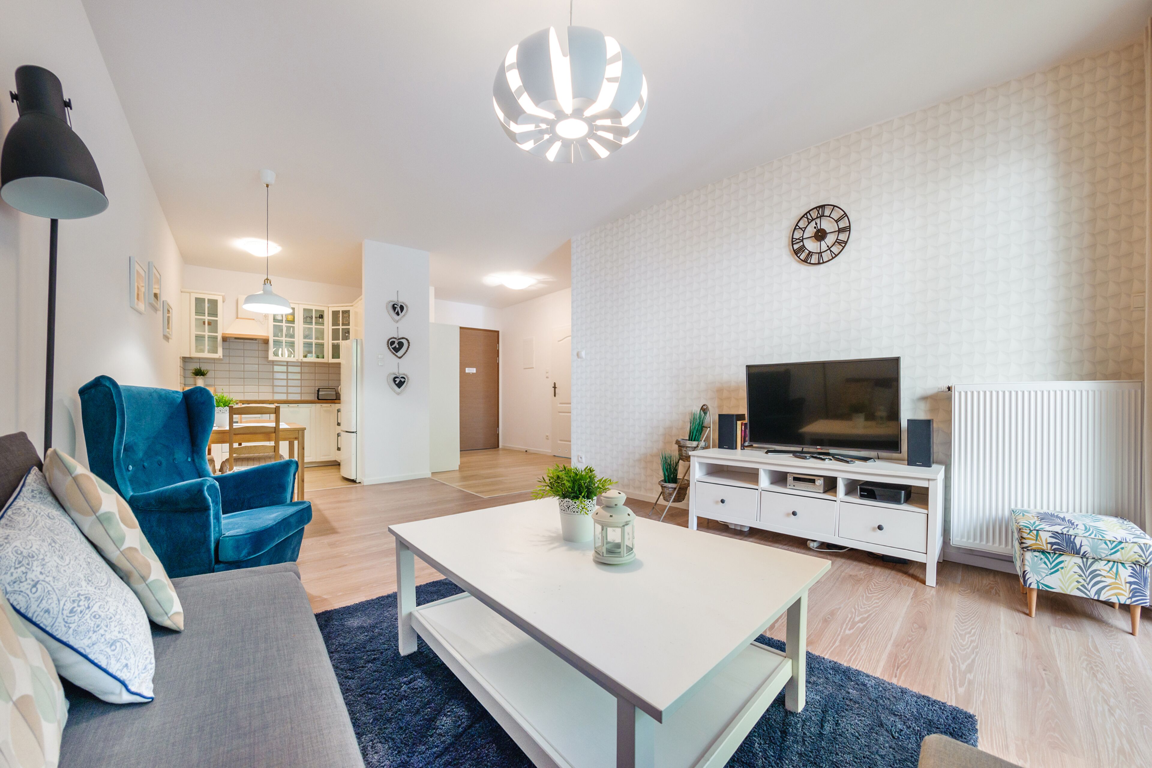 A modern living room with a sofa, a blue armchair, a white coffee table, a TV, and a kitchen in the background.