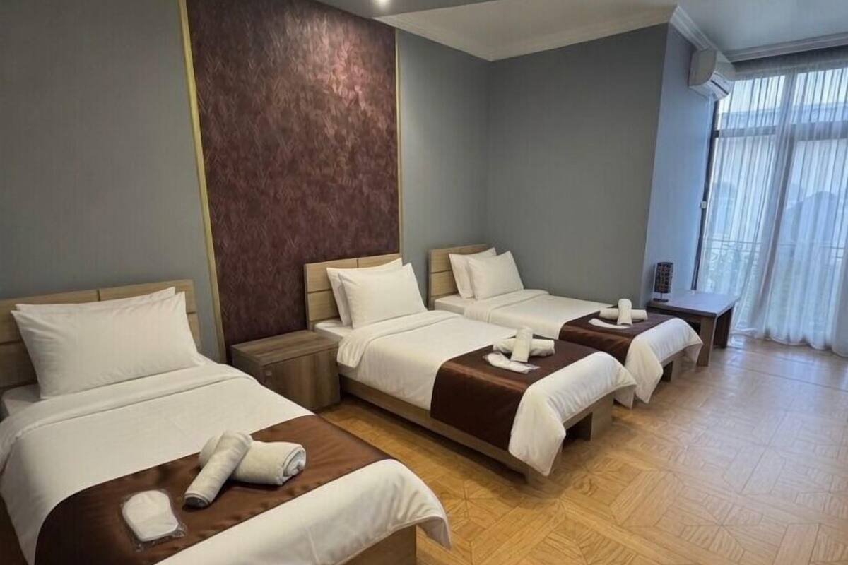 Ein Hotelzimmer mit drei Betten, Holzboden und einem großen Fenster mit Vorhängen.