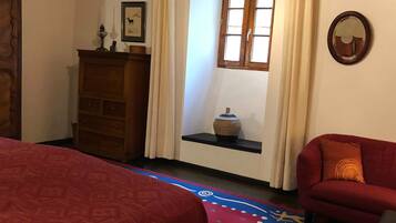 Quarto com cômoda de madeira, uma cadeira vermelha, tapete estampado e janela com cortinas.