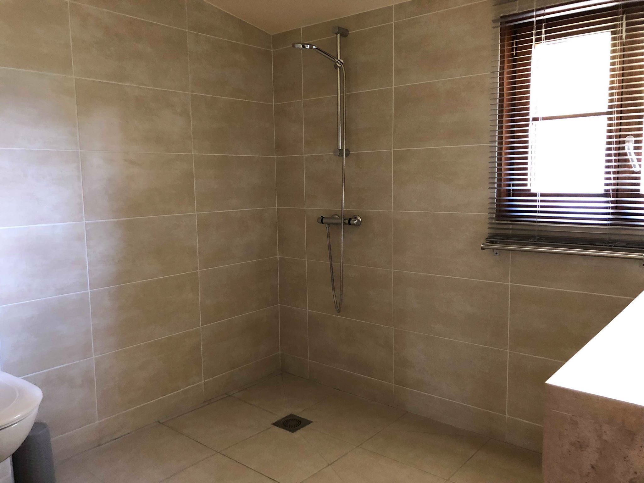 A bathroom with beige tiles, a shower, a window with blinds, and a toilet.