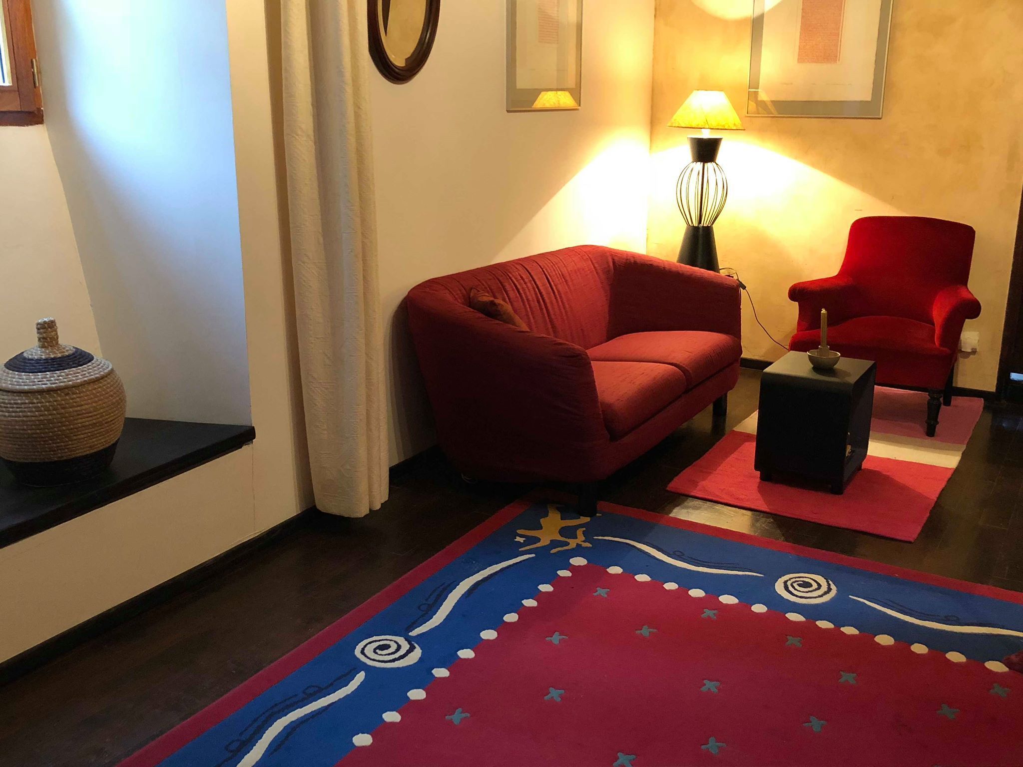 En stue med en rød sofa, en rød lænestol, et lille bord, en lampe og en dekorativ måtte.